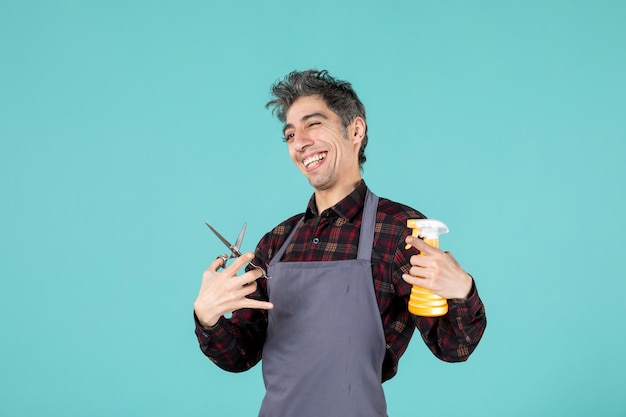 Vue de dessus d'un coiffeur souriant portant un tablier gris et tenant un vaporisateur de ciseaux sur une surface de couleur bleu pastel