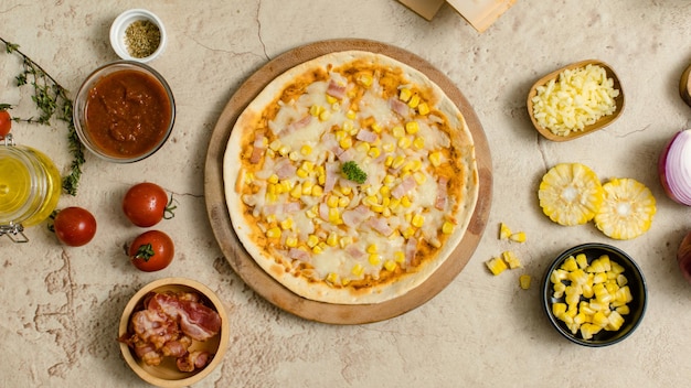 Vue de dessus d'une délicieuse pizza italienne au maïs et au jambon au fromage juteux et savoureuse placée sur une planche de bois sur la table de fête autour avec d'autres ingrédients sauce huile d'olive bacon tomates fromage et légumes