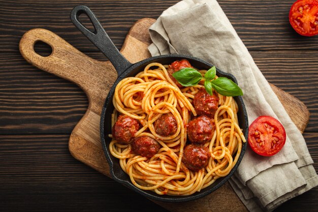 Vue de dessus de délicieuses pâtes aux boulettes de viande, sauce tomate et basilic frais dans une poêle vintage rustique en fonte servie sur une planche à découper, fond en bois. Savoureux spaghettis aux boulettes de viande maison