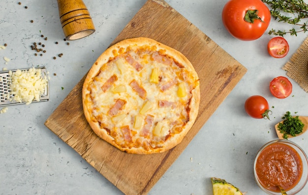 Vue de dessus de délicieux savoureux juteux de croûte italienne traditionnelle hawaïenne fine pizza croustillante au jambon et à l'ananas placé sur une planche à découper en bois avec des ingrédients tranches de tomates ketchup et sel