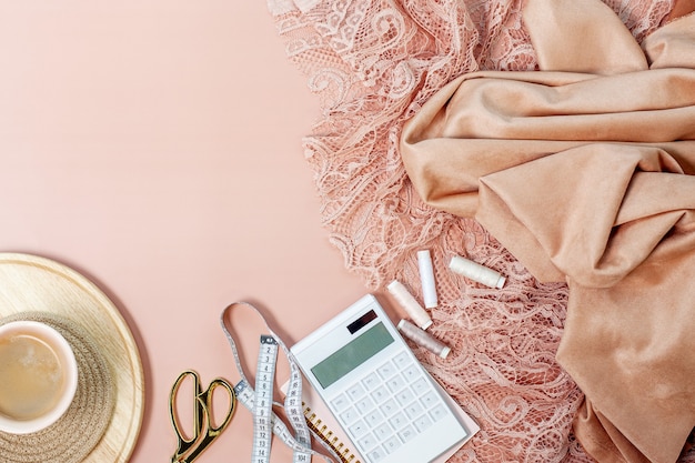 Vue de dessus de la dentelle de couleur corail rose et des tissus de soie avec des bobines de fil, calculatrice sur bloc-notes, ruban à mesurer, ciseaux de couture près de tasse de café sur fond rose. Concept de couture de robes de soirée.