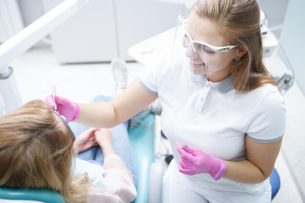 Vue de dessus d'un dentiste professionnel souriant, portant un écran facial en plastique tout en travaillant avec un patient