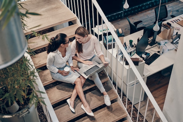 Vue de dessus de deux jeunes femmes modernes en vêtements décontractés intelligents communiquant et utilisant un ordinateur portable assis dans les escaliers