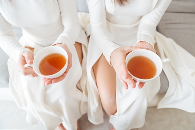 Vue de dessus de deux jolies femmes en robes blanches assises et tenant deux tasses de thé