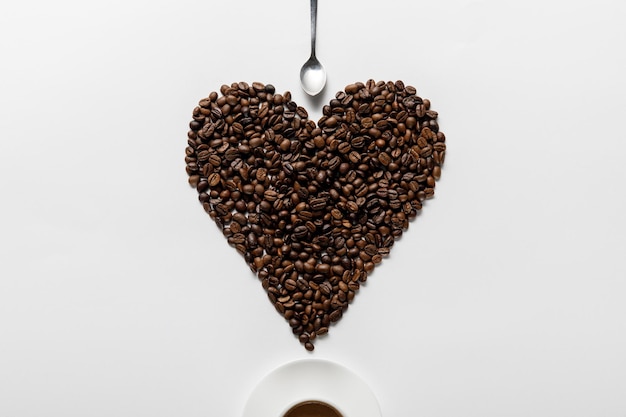 Vue de dessus du café dans une tasse sur une soucoupe avec un cœur fait de grains de café près d'une cuillère sur fond blanc