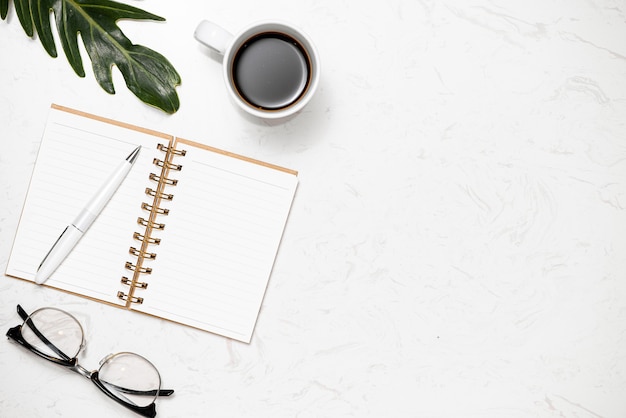 Vue de dessus du cahier ouvert pour notes, stylo, verres et tasse de café sur fond de marbre blanc.