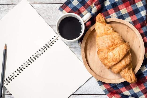 Vue de dessus du cahier vierge avec un crayon, croissant et café sur fond de table en bois.