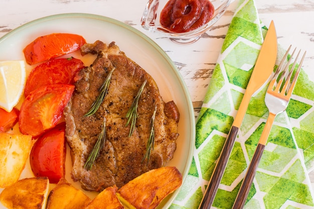 Vue de dessus du cou de porc grillé servi avec pommes de terre frites, tomates et cetcup sur fond clair.