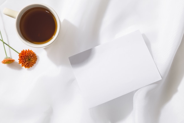 Vue de dessus du fond de tissu de voile avec une tasse de thé, deux fleurs orange et un morceau de papier blanc avec un espace pour le texte. Horizontal