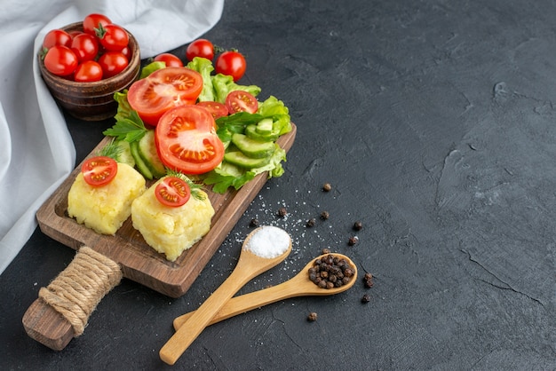 Vue de dessus du fromage entier de tomates fraîches et concombres coupés sur la coutellerie de planche de bois set épices dans des cuillères sur le côté droit sur la surface noire