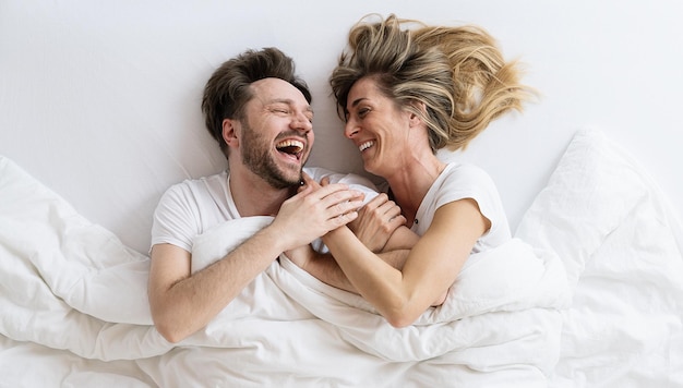 Vue de dessus du jeune couple d'amoureux allongé sur le lit en riant et en souriant avec une couverture. Ils se regardent avec confiance dans l'amour