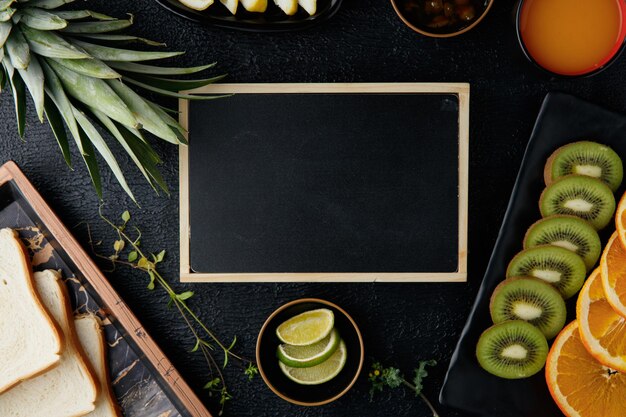 Photo vue de dessus du mini tableau noir avec kiwi orange citron tranches de citron vert feuilles d'ananas confiture jus d'orange et tranches de pain dans un plateau sur fond noir avec espace de copie