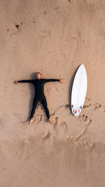 Photo vue de dessus du surfeur en combinaison avec planche de surf allongé sur la plage de sable
