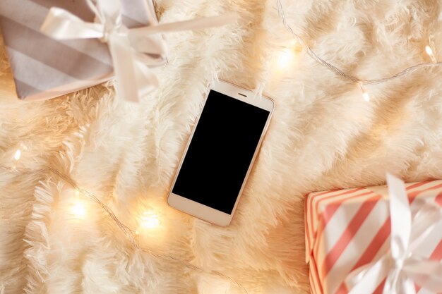 Vue de dessus du téléphone portable avec écran noir vierge pour la publicité ou le texte promotionnel près des boîtes de cadeaux de Noël sur un tapis blanc doux au sol avec des guirlandes lumineuses.