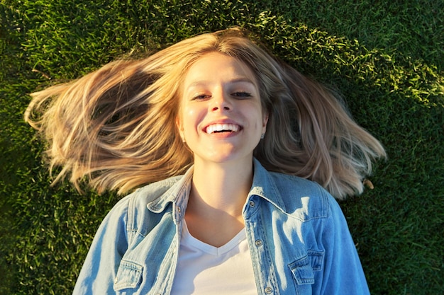 Vue de dessus du visage souriant et heureux de la belle jeune femme allongée sur l'herbe verte, journée ensoleillée. Émotions bonheur, joie, rêverie, plaisir