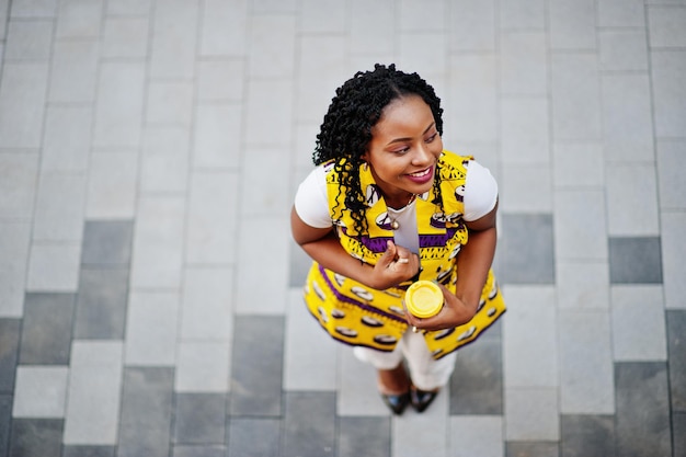 Vue de dessus d'élégantes femmes afro-américaines en veste jaune posées dans la rue avec une boisson chaude dans une tasse en papier jetable