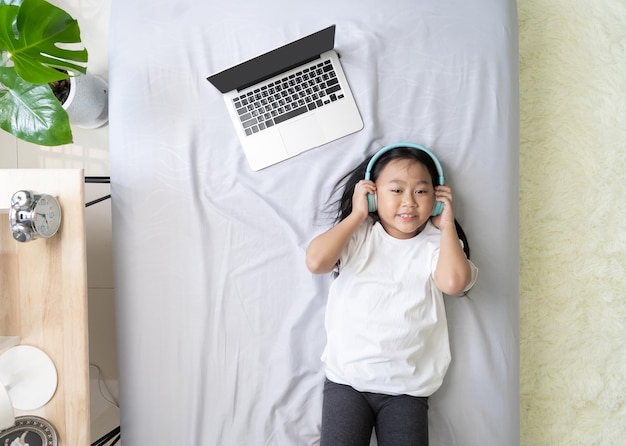 Vue de dessus Enfant asiatique petite fille portant des écouteurs avec un ordinateur portable sur le lit