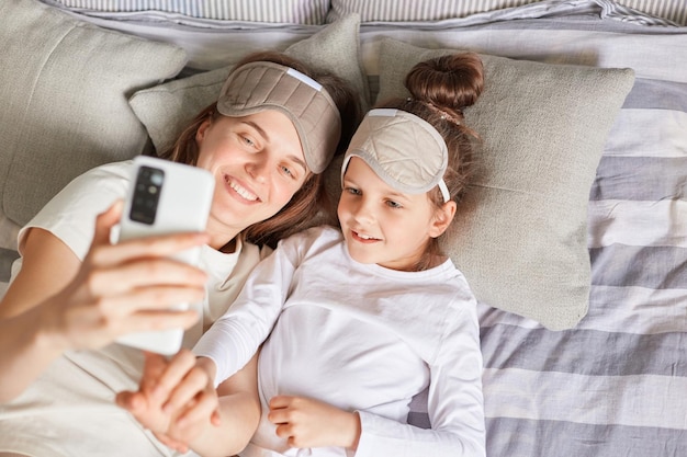 Vue de dessus d'une famille heureuse souriante se réveillant le matin allongé dans son lit dans des masques de sommeil et faisant selfie femme tenant un téléphone intelligent dans les mains prenant une photo