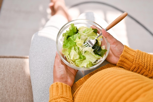 Vue de dessus d'une femme enceinte tenant un saladier mangeant de la salade sur un canapé dans son salon