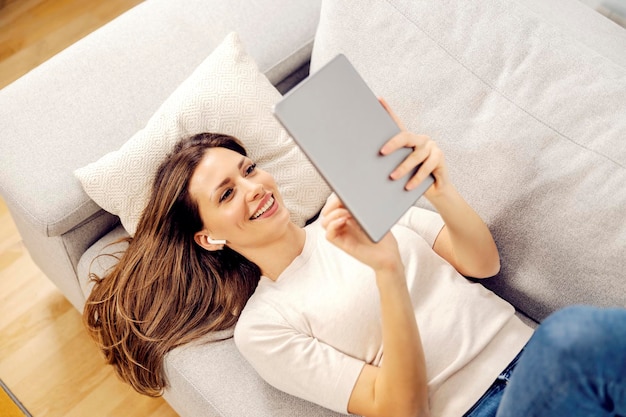 Vue de dessus d'une femme heureuse allongée sur un canapé dans sa maison confortable et utilisant une tablette