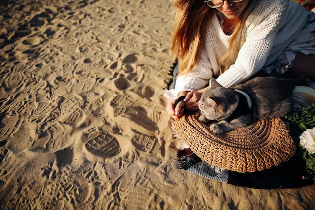 Vue de dessus d'une femme sur la plage avec son chaton gris rectangulaire écossais de race au coucher du soleil Le chat se trouve sur un sac de paille près des fleurs