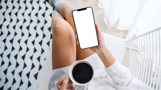 Vue de dessus d'une femme tenant un téléphone mobile avec un écran de bureau blanc vierge tout en étant assis et en buvant du café dans la chambre