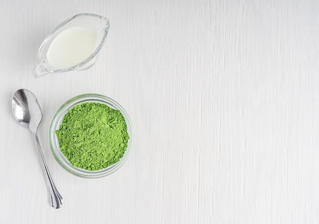 Vue de dessus des feuilles de thé vert matcha en poudre moulue avec du lait végétal et une cuillère sur une table en bois blanc