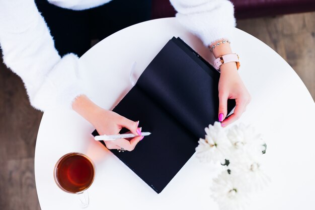 Vue de dessus d'une fille dans un café qui est assis à la table et écrit dans un cahier avec des pages noires