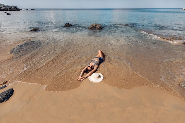 Vue de dessus d'une fille mince en train de bronzer sur une plage de sable ensoleillée pendant des vacances en mer. A proximité se trouve un chapeau de paille. Les vagues de la mer touchent ses jambes.