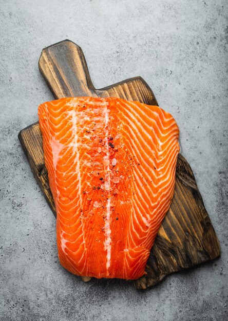 Vue de dessus, gros plan de filet de saumon cru entier frais avec assaisonnements sur planche de bois, fond de pierre grise. Préparation du filet de saumon pour la cuisson, concept d'alimentation saine