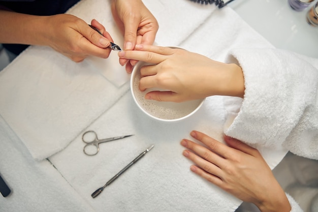 Vue de dessus en gros plan de la main féminine au-dessus du bain de manucure pendant que le professionnel traite les ongles avec une pince