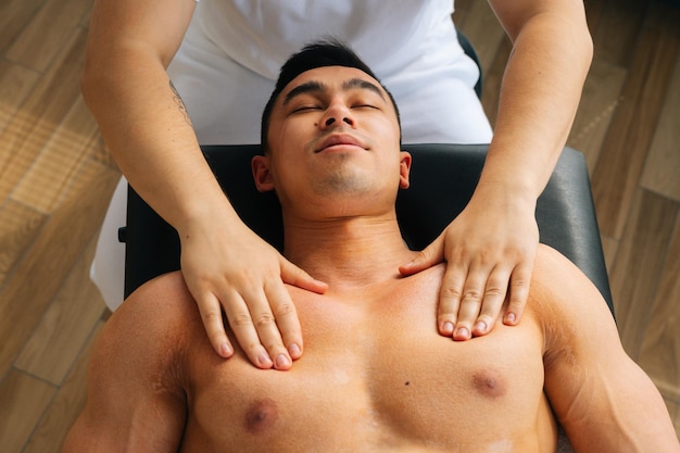 Vue de dessus en gros plan d'un masseur masculin professionnel avec des mains fortes massant les muscles des épaules et de la poitrine d'un athlète musclé après une séance d'entraînement physique, allongé sur le dos dans une table de massage.