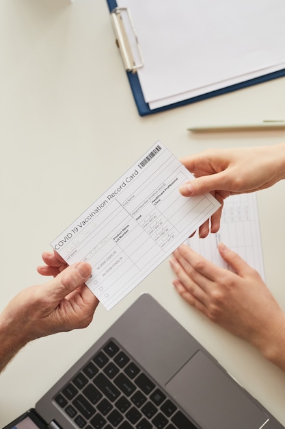 Vue de dessus en gros plan d'un médecin donnant une carte de vaccination Covid au patient de l'autre côté de la table dans la clinique, espace de copie