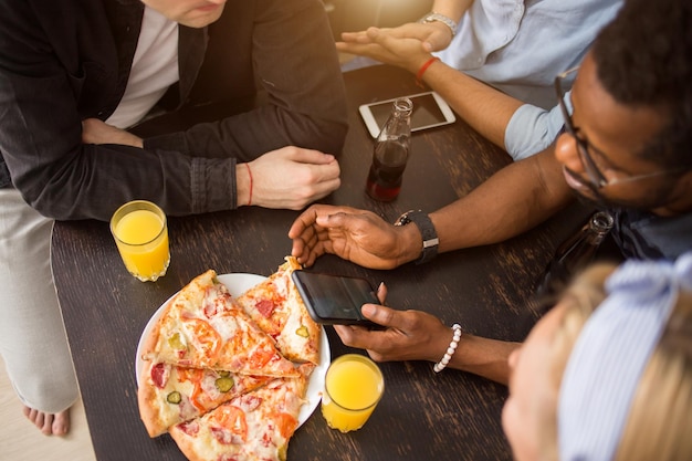 Vue de dessus d'un groupe d'amis assis à table dans un café, déjeunant, mangeant de la pizza, buvant du jus, utilisant un téléphone portable, écrivant une critique sur les réseaux sociaux. Livraison, nourriture, concept technologique.