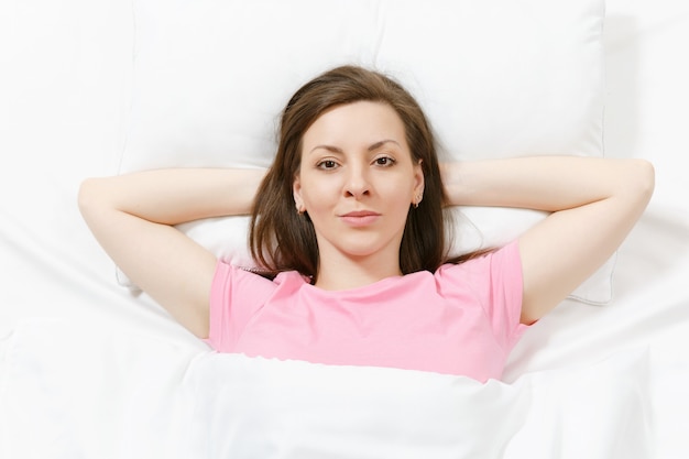 Vue de dessus de l'heureuse jeune femme brune allongée dans son lit avec un drap blanc, un oreiller, une couverture, les mains sous la tête. Femme souriante passant du temps dans la chambre