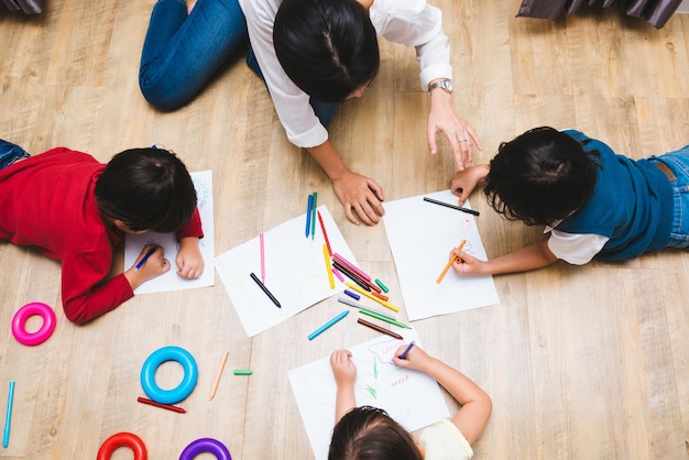 Vue de dessus heureux famille asiatique enfants groupe enfant garçon et fille maternelle jouant peinture dessin couleur sur papier avec formation des enseignants ensemble à la salle de jeux intérieure, concept de retour à l'école