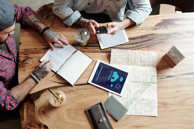 Vue de dessus de l'homme tatoué à prendre des notes dans le planificateur tout en se préparant à voyager avec un ami au café