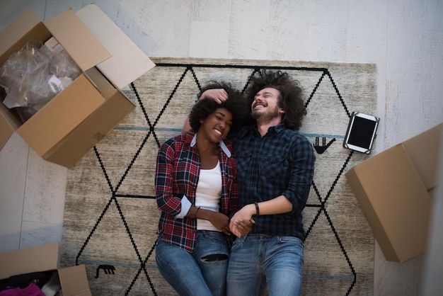 Photo vue de dessus d'un jeune couple multiethnique séduisant se déplaçant, se tenant la main, regardant la caméra et souriant en position couchée parmi des boîtes en carton