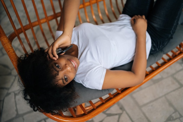 Vue de dessus d'une jeune femme afro-américaine joyeuse parlant sur un smartphone allongé sur un canapé confortable ayant une conversation téléphonique agréable dans le salon à la maison