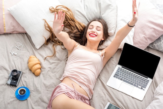 Vue de dessus sur la jeune femme allongée sur le lit avec ordinateur portable, téléphone et appareil photo. Repos après le travail