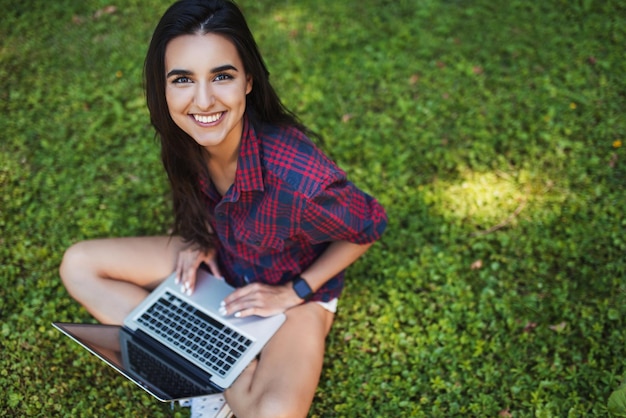 Vue de dessus d'une jeune femme brune caucasienne souriante et portant une chemise à carreaux assise sur l'herbe devant un ordinateur portable travaillant sur son ordinateur portable avec une communication sans fil faisant du shopping et étudiant