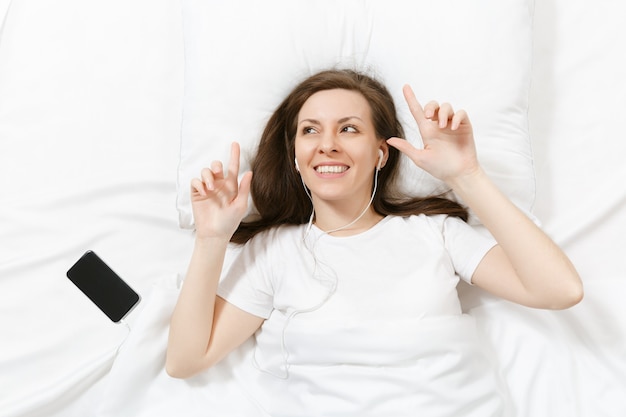 Vue de dessus d'une jeune femme heureuse allongée dans son lit avec un drap blanc, un oreiller, une couverture, écouter de la musique à partir d'écouteurs dans un téléphone portable