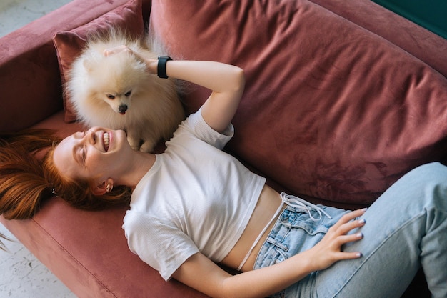 Vue de dessus d'un joli petit chien Spitz blanc adorable léchant le visage d'une jeune femme riante allongée sur un canapé confortable