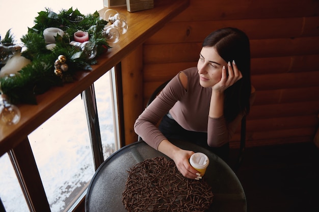 Vue de dessus d'une jolie femme tenant une tasse de papier à emporter avec boisson chaude, assise au café en bois et regardant par la fenêtre. Belle décoration de Noël sur la planche de bois à côté de la fenêtre