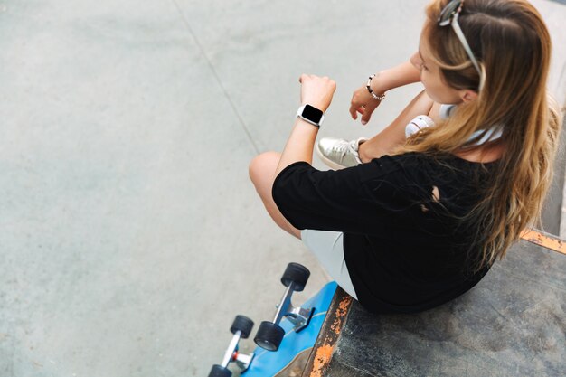 Vue de dessus d'une jolie jeune adolescente assise au skatepark avec un longboard, vérifiant sa montre intelligente