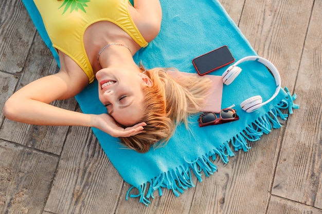 Vue de dessus d'une jolie jeune fille blonde souriante portant un maillot de bain relaxant sur une couverture d'été à la plage sur du parquet