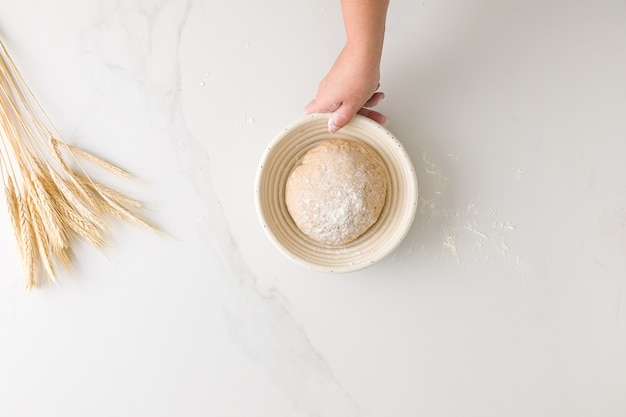 Vue de dessus de la main féminine tenant une pâte à pain au repos dans un bol à pain dans une table en marbre avec du blé et de la farine avec un espace pour le texte