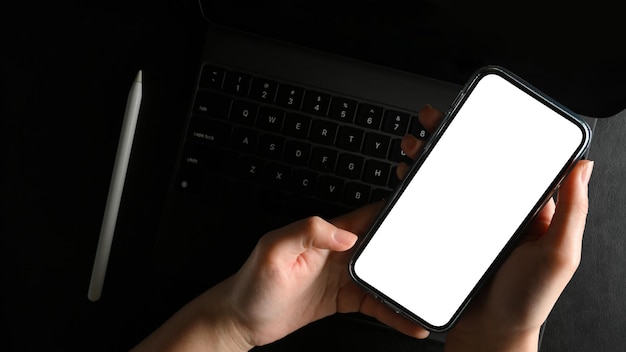 Vue de dessus d'une main de femme tenant une maquette d'écran vierge de smartphone sur un bureau de travail sombre et moderne