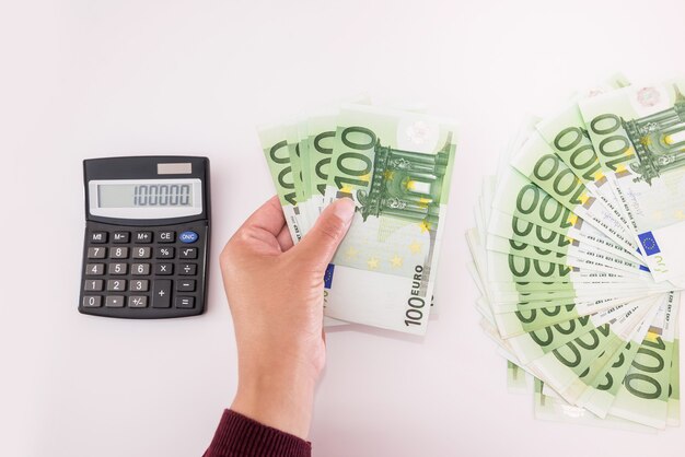 Vue de dessus des mains féminines avec des billets en euros et une calculatrice sur blanc. Notion financière.