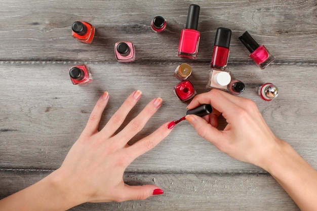 Vue de dessus sur les mains de la femme, appliquant une couche de vernis à ongles rouge sur ses ongles, avec beaucoup de vernis à ongles sur un bureau en bois gris en arrière-plan.